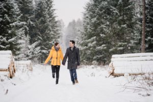 Ein Verlobungsbild. Ein Pärchen läuft hopsend durch eine Schneelandschaft. Links und rechts sind Bäume auf dem Boden gestapelt