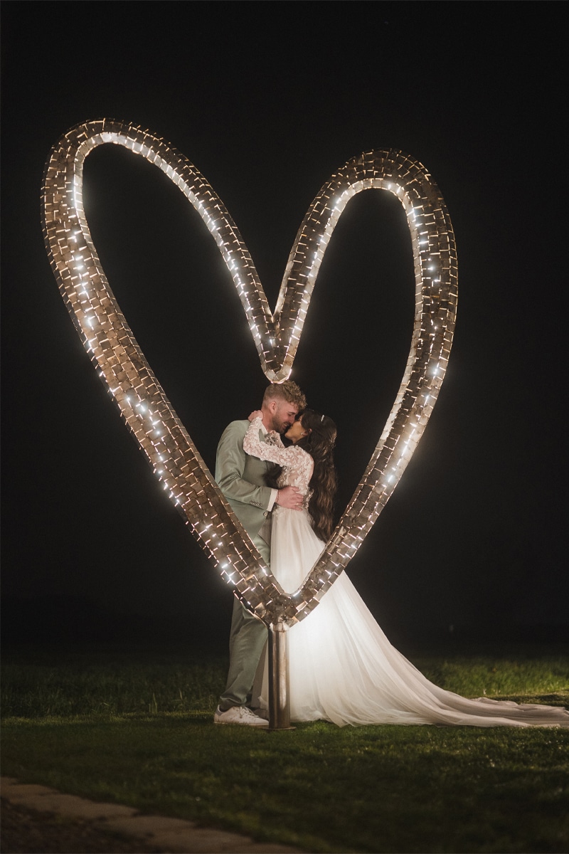 Ein Brautpaar nachts küsst sich und wird von einem Herz aus Metall mit Lichtern angestrahlt
